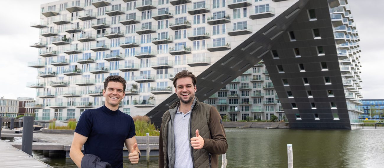 Twee mannen staan glimlachend buiten voor een groot, modern gebouw met veel balkons en een opvallende diagonale constructie; beide tonen een duim omhoog.
