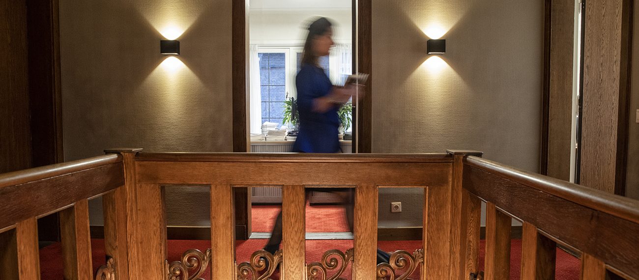 Interieur met houten balustrade, warme wandverlichting en passerende persoon in klassieke gang.