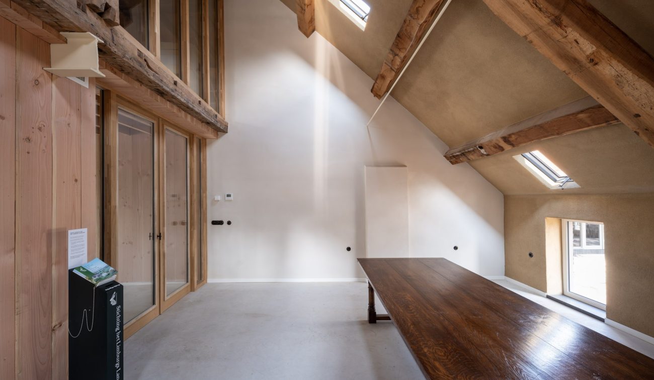 Gerestaureerde zolderruimte van de Grathemermolen met zichtbare houten spanten, wit gestucte wanden, dakramen en een lange massiefhouten tafel naast een glazen scheidingswand.