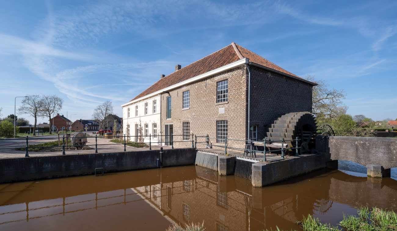 Grathemermolen in Grathem – bakstenen watermolen met rood pannendak en groot waterrad aan het water, met spiegeling in de molenvijver onder blauwe lucht.