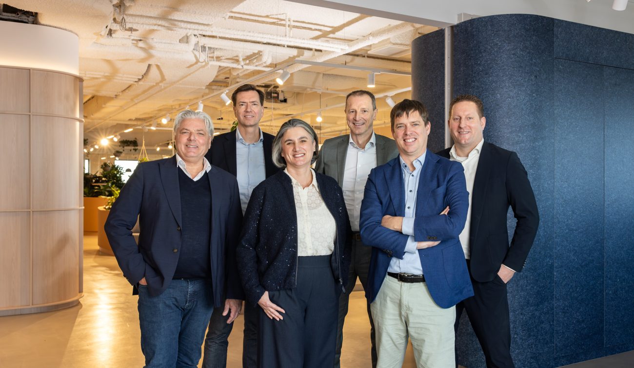 Groep van zes netjes geklede volwassenen die samen poseren in een moderne kantooromgeving, met planten en warme verlichting op de achtergrond.