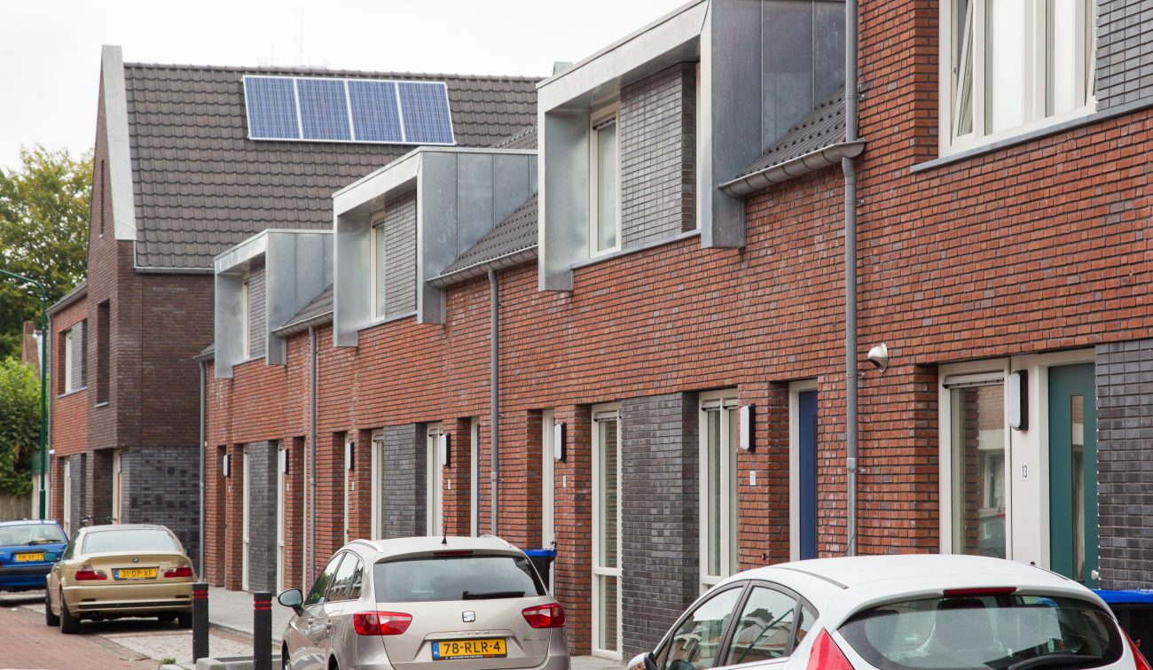 Nieuwbouwwoningen in Zevenbergen met moderne baksteenarchitectuur, rode en donkergrijze gevels, dakkapellen met zinken bekleding en zonnepanelen op het dak, met geparkeerde auto’s in de straat.