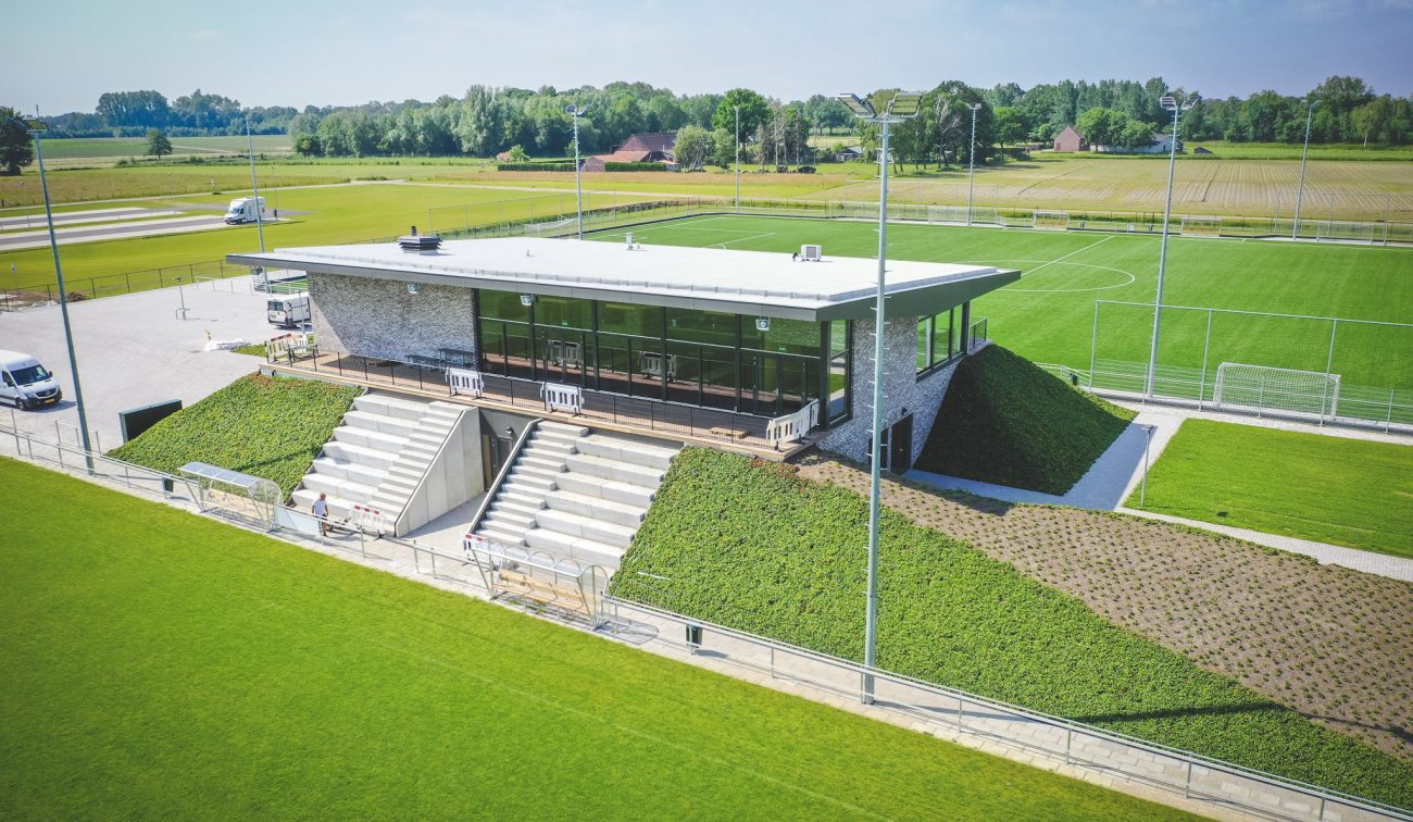 Luchtfoto van het moderne clubgebouw van DVO’20 in Kelpen-Oler, met glazen pui, tribunetrap en groene taluds, omgeven door meerdere voetbalvelden.