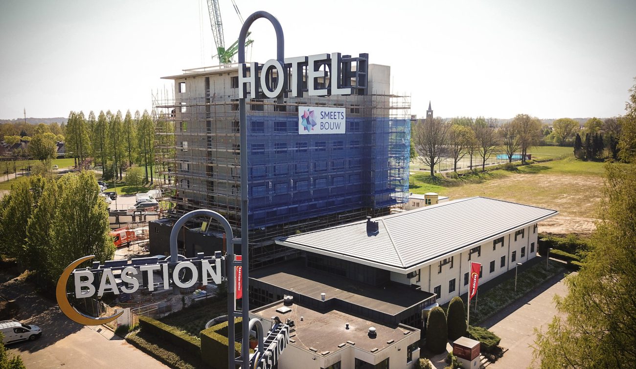 Bouwfase Bastion Hotel Geleen: hoge hotel­toren in steigers met blauw net en kraan, banner “Smeets Bouw” aan de gevel; op de voorgrond de grote “BASTION HOTEL”-lichtreclame en rechts het lagere bestaande hotelgebouw met grijs zadeldak, omringd door groen.