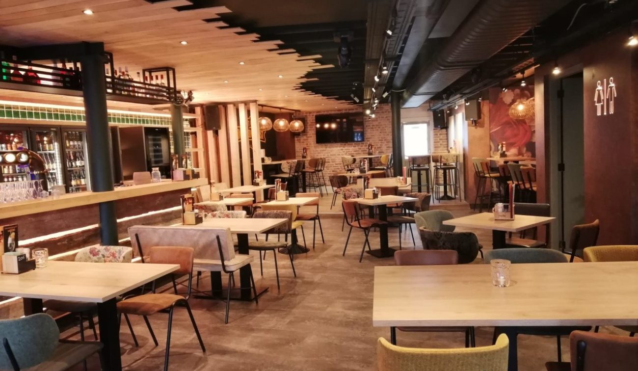 ChatGPT zei:  Interieur van eetcafé De Aanwinst in Nederweert: warme zithoek met houten tafels en diverse stoelen, lange bar met flessenkoelingen links, industriële zwarte luchtkanalen aan het plafond en toiletpictogram aan de rechterwand.