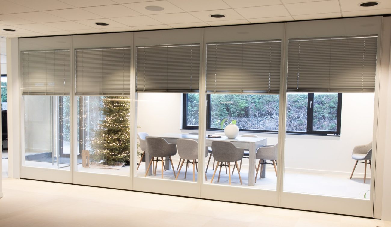 Vergaderruimte achter een glazen wand met jaloezieën: grijze stoelen rond een lichte tafel met witte vaas, raam met zicht op heg; links een verlichte kerstboom in de hal.