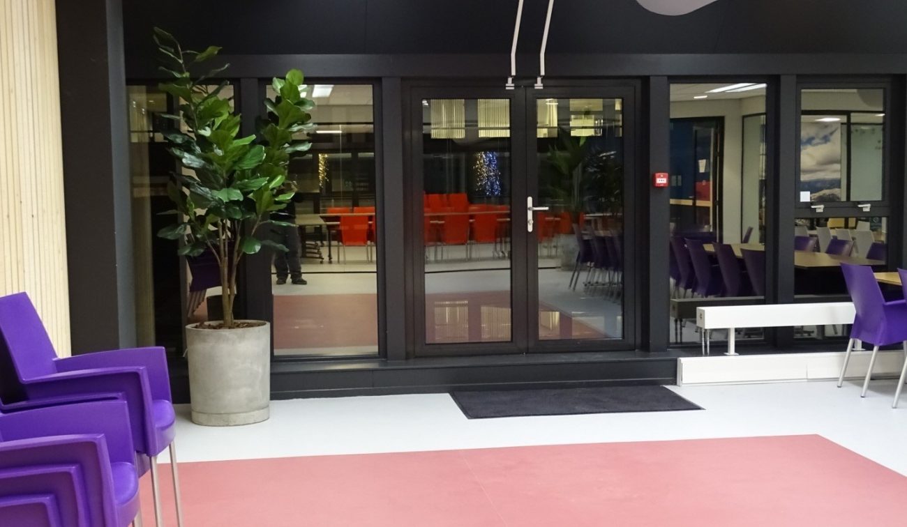 Entreezone Meeting Plaza: zwarte glaspuien met deur, plant in pot, mat en zicht op oranje stoelen