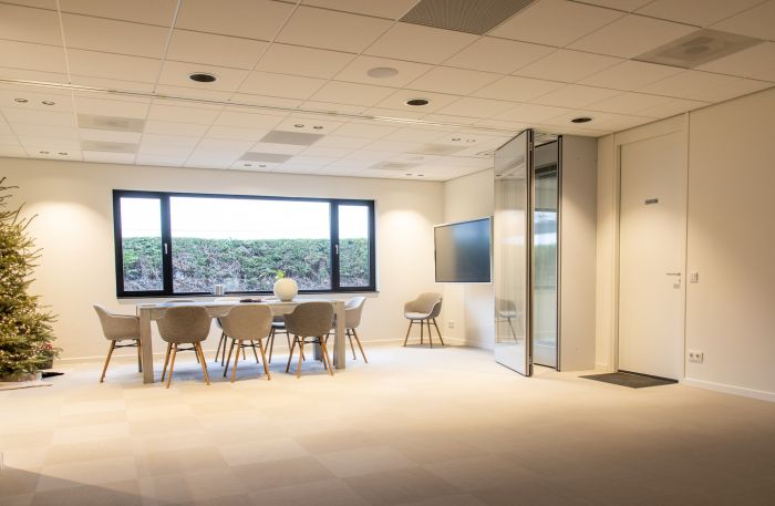 Lichte vergaderruimte met lange tafel en zes grijze stoelen bij een groot raam; rechts een wandmonitor, een opgevouwen glazen/akoestische scheidingswand en een witte deur.