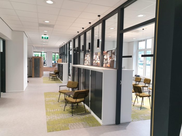 Lichte kantoorruimte met gang en glazen scheidingswanden in zwarte frames; hanglampen langs het plafond, groene fauteuils op tapijttegels en fotopanelen op witte kasten. Grote ramen zorgen voor daglicht.