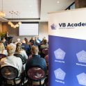 Een groep mensen zit op stoelen in een zaal naar een presentator te luisteren, in de hoek staat een roll up banner met de tekst "VB Academy" er op