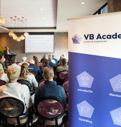 Een groep mensen zit op stoelen in een zaal naar een presentator te luisteren, in de hoek staat een roll up banner met de tekst "VB Academy" er op