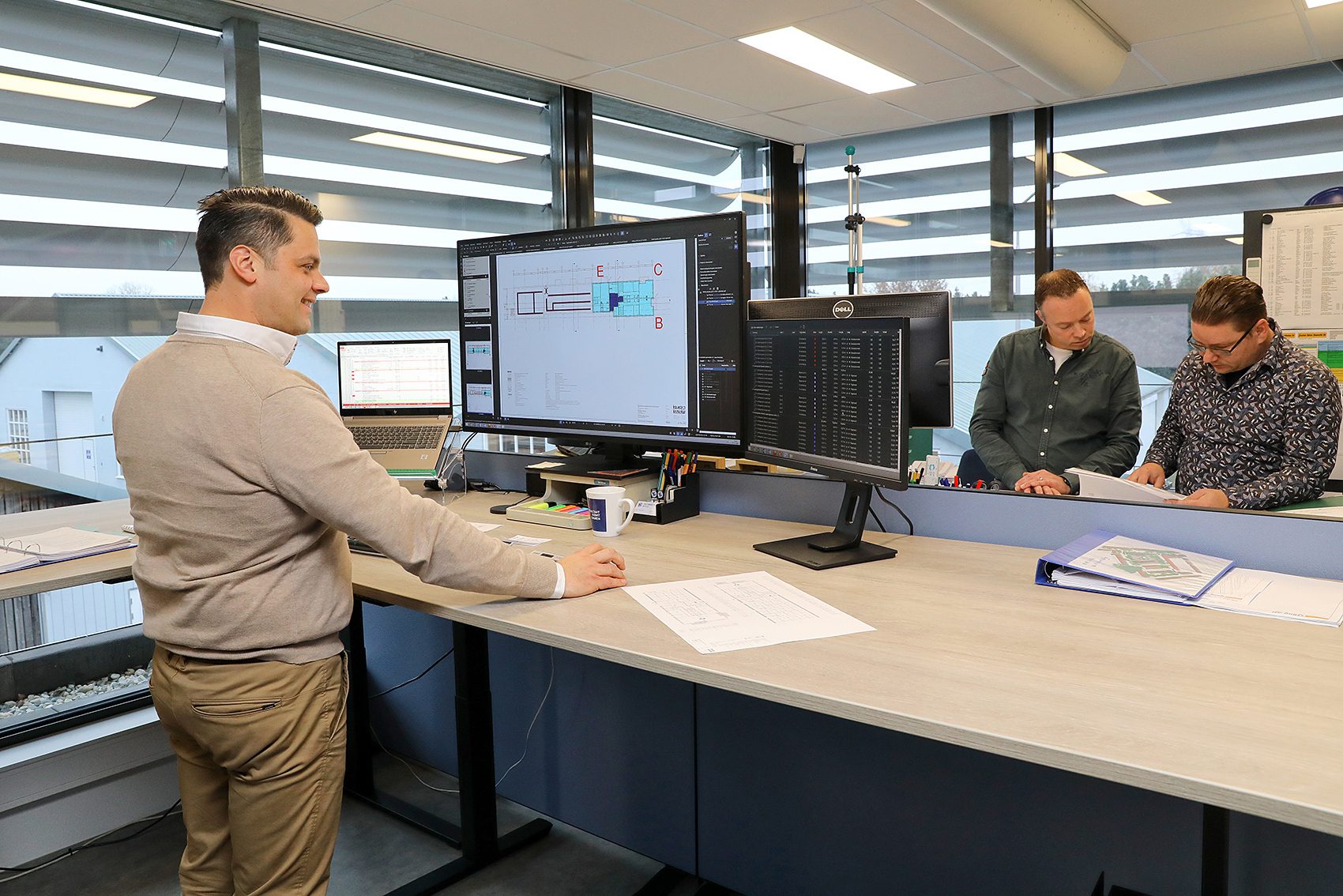Medewerker bekijkt bouwtekeningen op groot scherm in kantoor terwijl twee collega’s samen technische documenten doornemen.