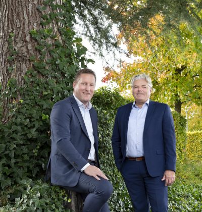 Twee mannen in pak poseren buiten bij een boom en groen struikgewas.