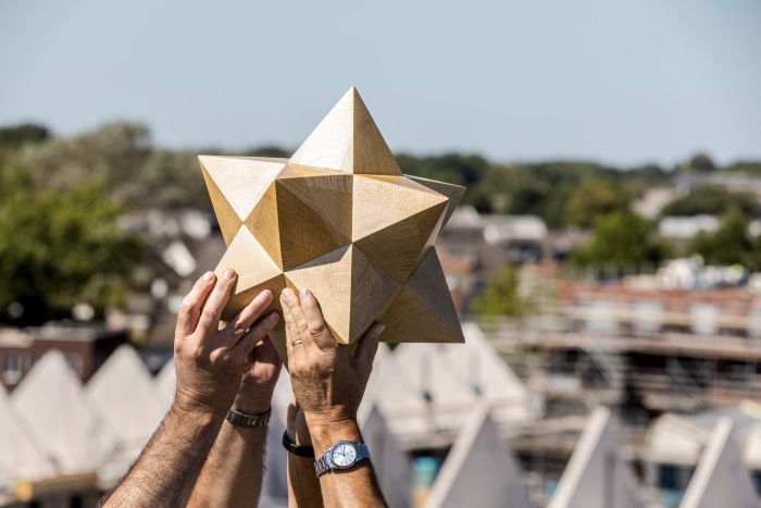 Verschillende handen tillen een houten diamant in de lucht, met op de achtergrond een zonnig verlichte bouwplaats
