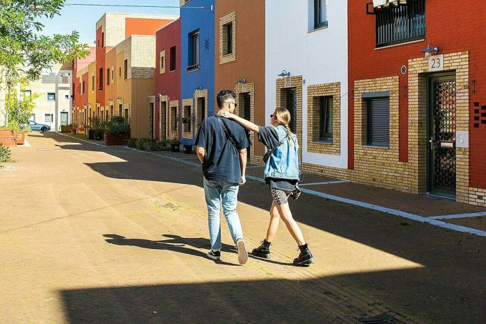 Twee mensen lopen in de zon door een straat met kleurrijke huizen op de achtergrond