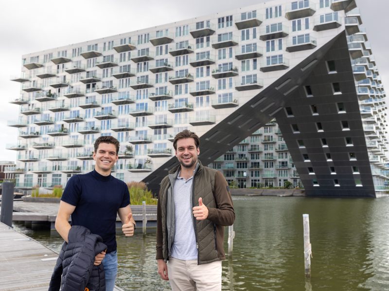 Twee mannen staan glimlachend buiten voor een groot, modern gebouw met veel balkons en een opvallende diagonale constructie; beide tonen een duim omhoog.