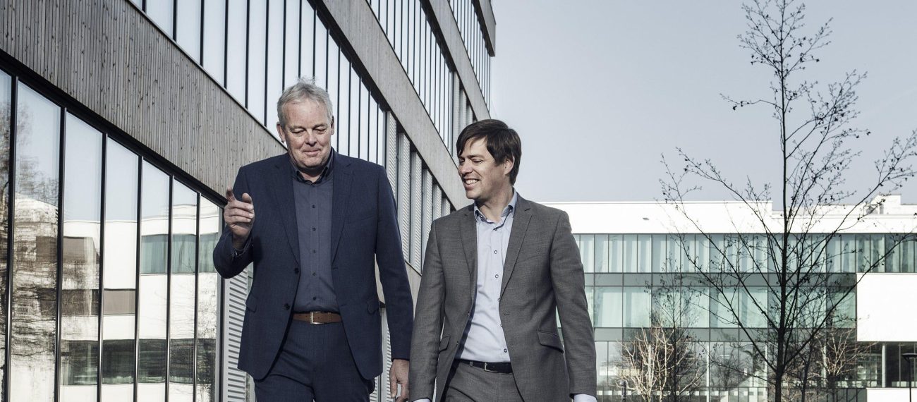 Twee mannen in pak lopen en praten samen langs een modern kantoorgebouw.
