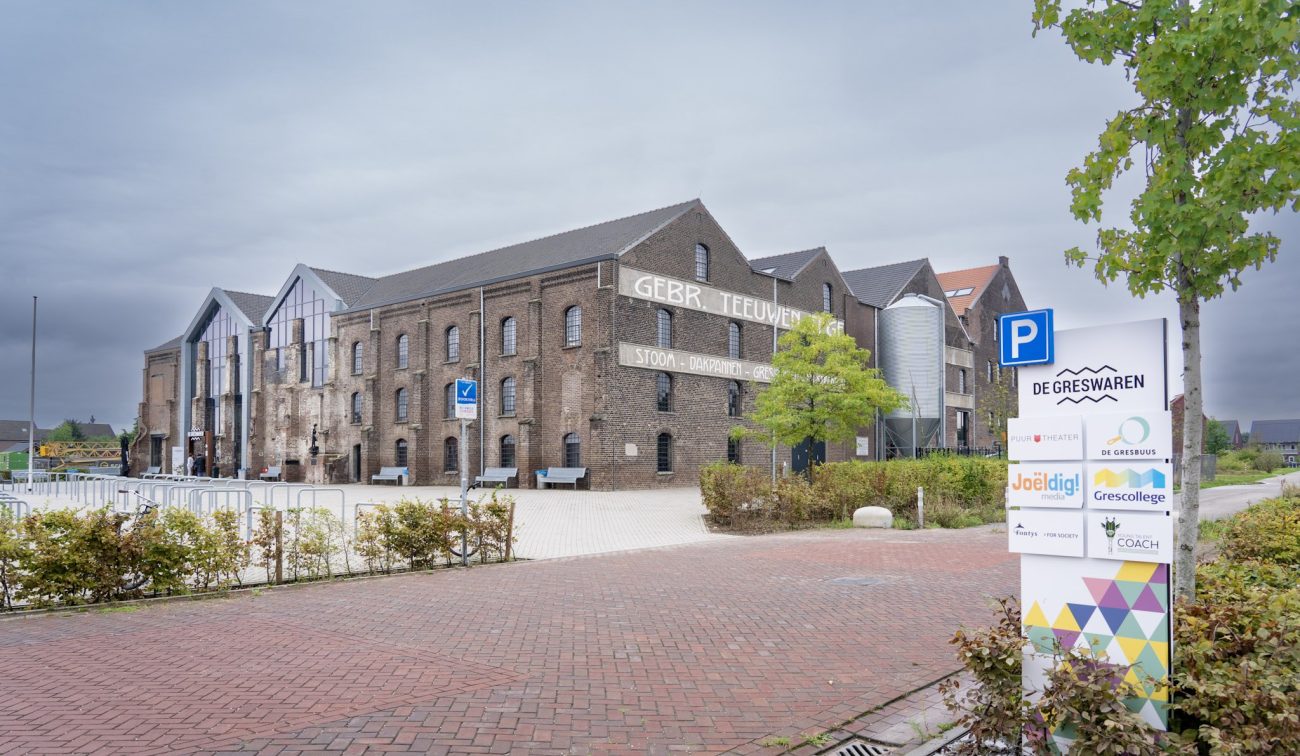 Gerestaureerde Greswarenfabriek in Reuver met bakstenen gevel, grote glazen entree en historische opdruk ‘GEBR. TEEUWEN’; voorplein met bankjes, fietsrekken en een jonge boom.