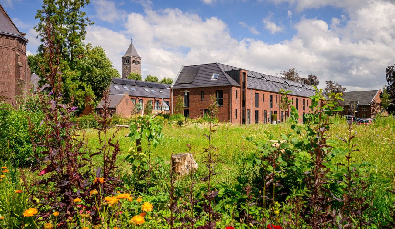 Gebiedsontwikkeling Kloostervelden in Sterksel met nieuwbouw rijwoningen in rood baksteen met zonnepanelen rond een groene, bloemrijke binnentuin en op de achtergrond een kloostertoren.