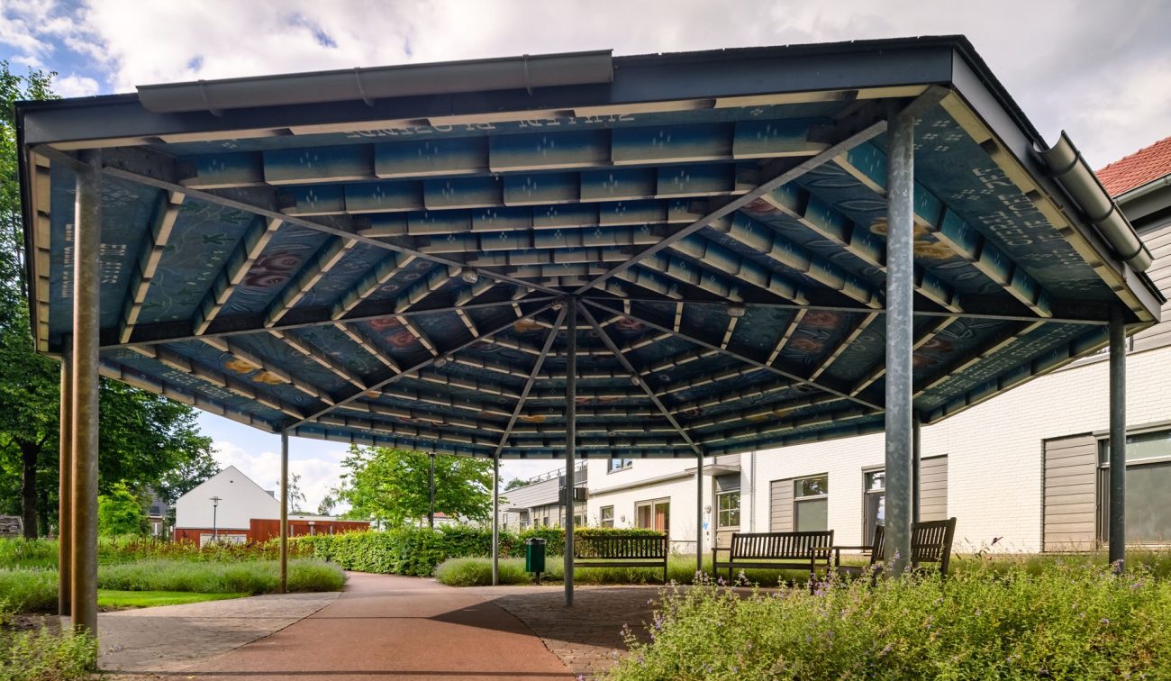 Overdekte ontmoetingsplek met kunstzinnig blauw dak, bankjes en groene wandelpaden tussen witte woningen in Kloostervelden, Sterksel.