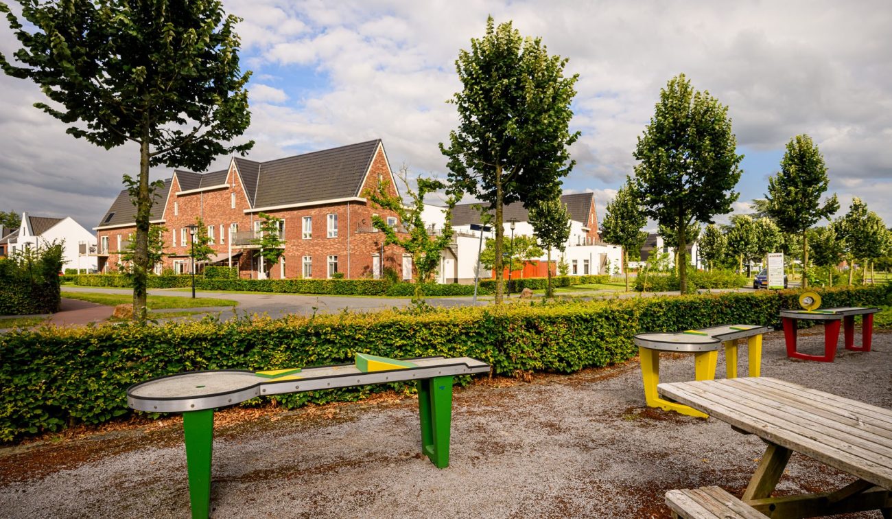 Speel- en zitplek met kleurrijke tafels aan groen plantsoen, met nieuwe bakstenen en witte woningen op de achtergrond in Kloostervelden, Sterksel.
