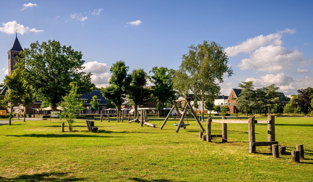 Groen speelveld met houten speeltoestellen in Kloostervelden, Sterksel, omringd door bomen en nieuwe woningen onder een zonnige blauwe lucht.