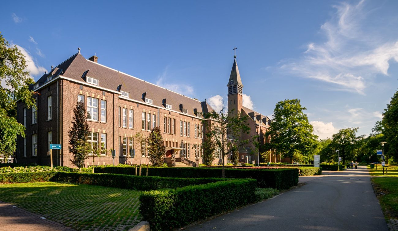 Historisch kloostercomplex met kerktoren in Kloostervelden, Sterksel, omgeven door strakke hagen en volwassen bomen op een zonnige dag met blauwe lucht.