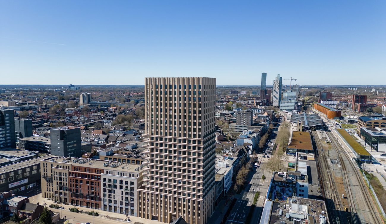 Ontwikkeling De Bankier Tilburg – luchtfoto van de woontoren langs het spoor, omringd door stedelijke bebouwing en het stationsgebied onder een heldere blauwe lucht.