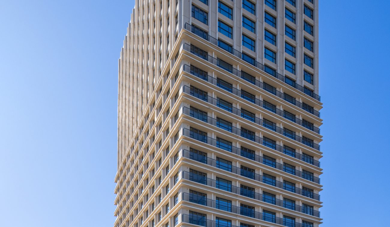 Ontwikkeling De Bankier Tilburg – volledig straatbeeld van de slanke woontoren met horizontale geveldetaillering en grote ramen, op de hoek bij het station onder een heldere blauwe lucht.