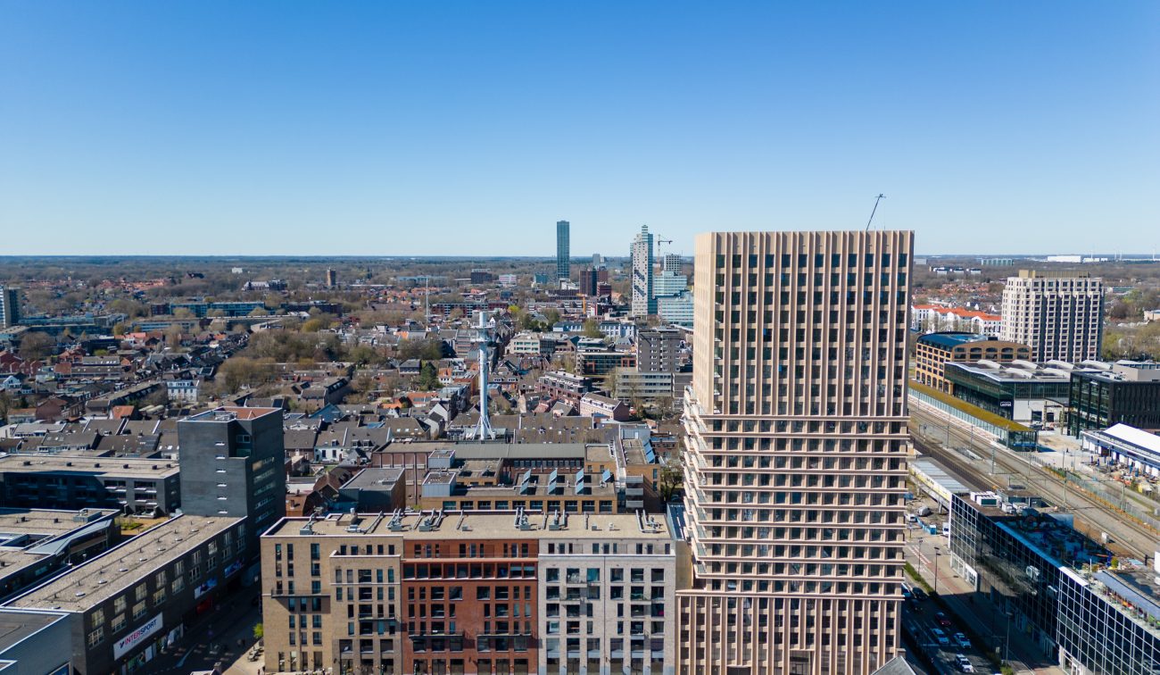 Ontwikkeling De Bankier Tilburg – luchtfoto met de nieuwe woontoren prominent in de skyline, naast het spoor en omliggende stedelijke bebouwing onder een heldere blauwe lucht.