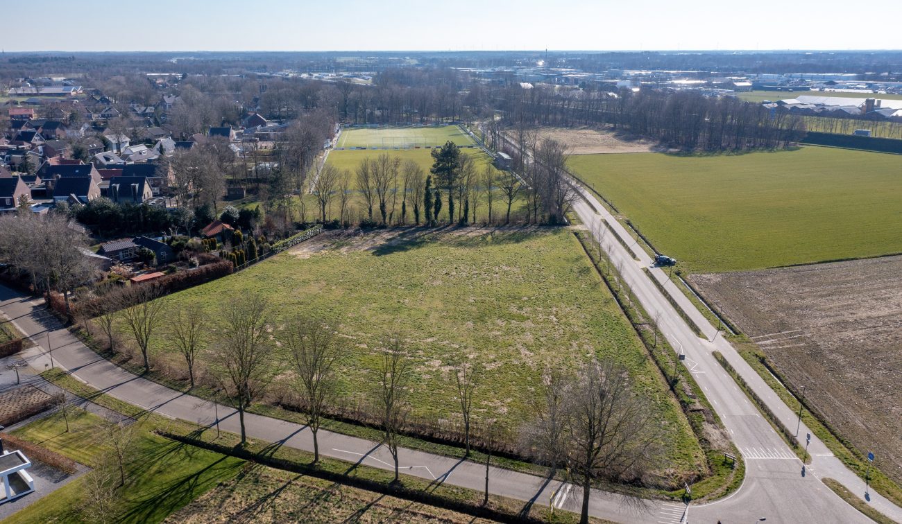 Vogelvlucht: groen bouwveld langs rechte ontsluitingsweg naast voetbalvelden en woonwijk — Ontwikkeling Kluisstraat Ommel