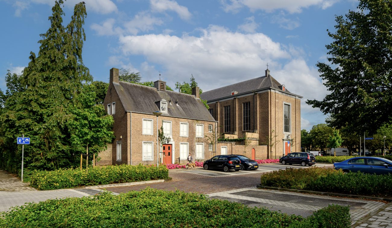 Mariakerk Valkenswaard – straatbeeld met voorhuis en kerk naast elkaar, parkeerplaatsen met auto’s en groene plantvakken onder een licht bewolkte lucht.