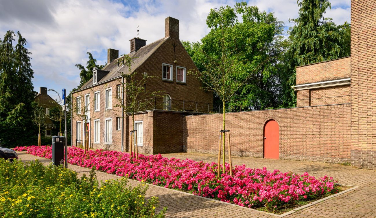 Transformatie Mariakerk Valkenswaard – voorhuis met bakstenen muur en rode poortdeur, brede stroken roze bloemperken en jonge bomen op een zonnige dag.