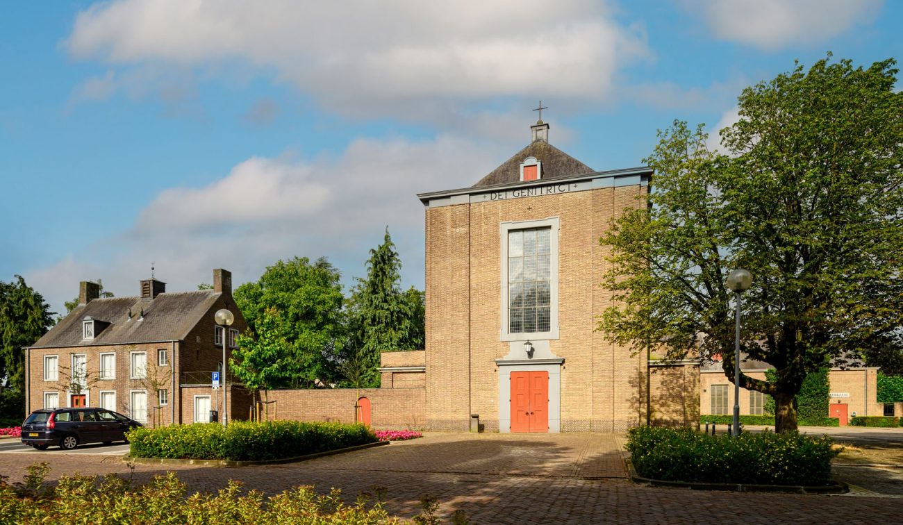 Transformatie Mariakerk Valkenswaard – vooraanzicht van de bakstenen kerk met rode deuren en hoge raamgevel, omringd door groen en pleintje onder blauwe lucht.
