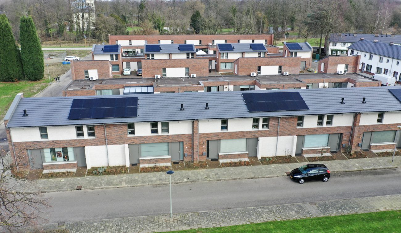 Oblique luchtfoto van nieuwe rijwoningen in Nazareth (Maastricht) met wit-bakstenen gevels en grijze zadeldaken met grote rijen zonnepanelen; daarachter meerdere blokken met platte daken. Een geparkeerde auto staat langs de straat.