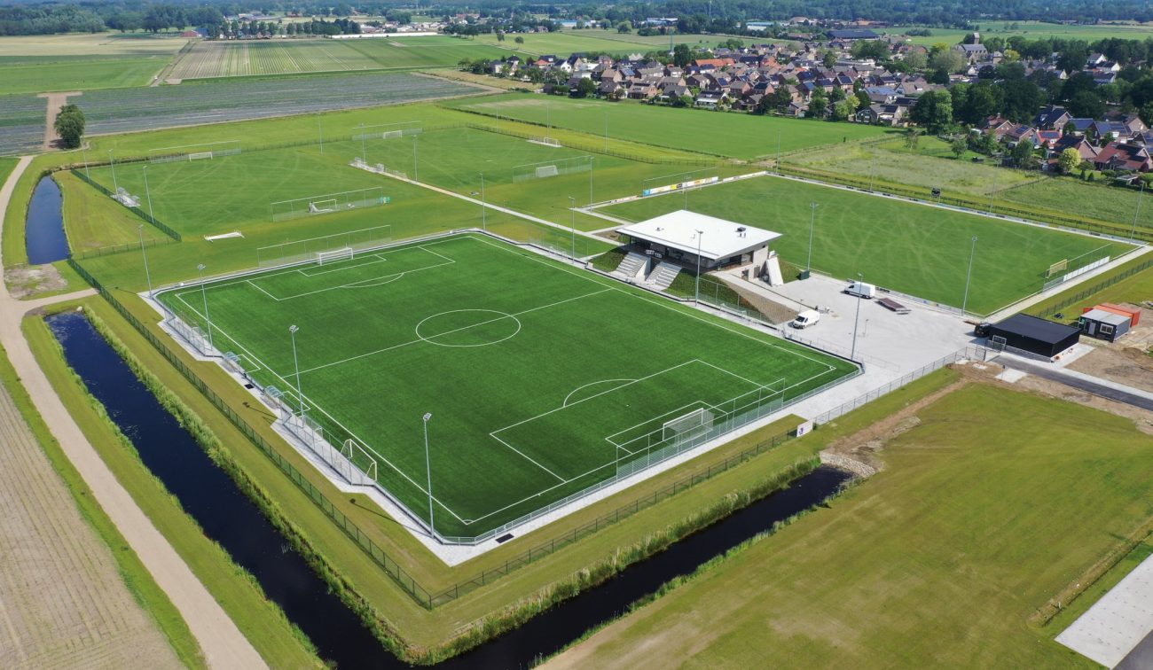 Luchtfoto sportpark DVO’20: hoofdveld met kunstgras, verlichtingsmasten en omheining, centraal het nieuwe clubgebouw met traptribune en groene taluds, omringd door extra velden, parkeerterrein en watergang.