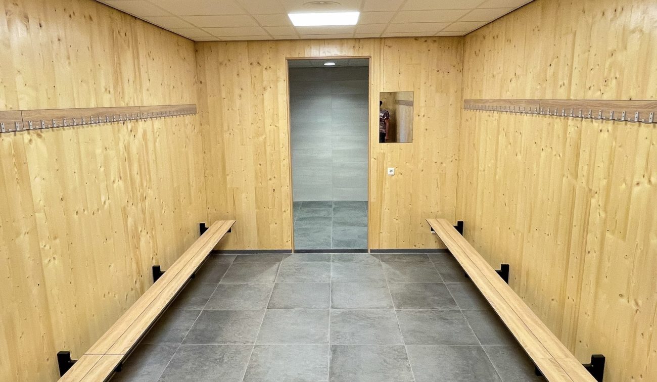 Kleedkamer in het nieuwe clubgebouw – houten wanden met kapstokhaken, lange bankjes langs beide zijden en een doorgang naar de doucheruimte.
