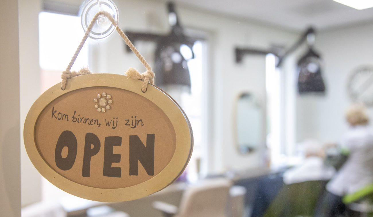 Ovaal bordje met tekst ‘Kom binnen, wij zijn OPEN’ aan een zuignap op de ruit, met een wazig salon/winkelinterieur op de achtergrond.
