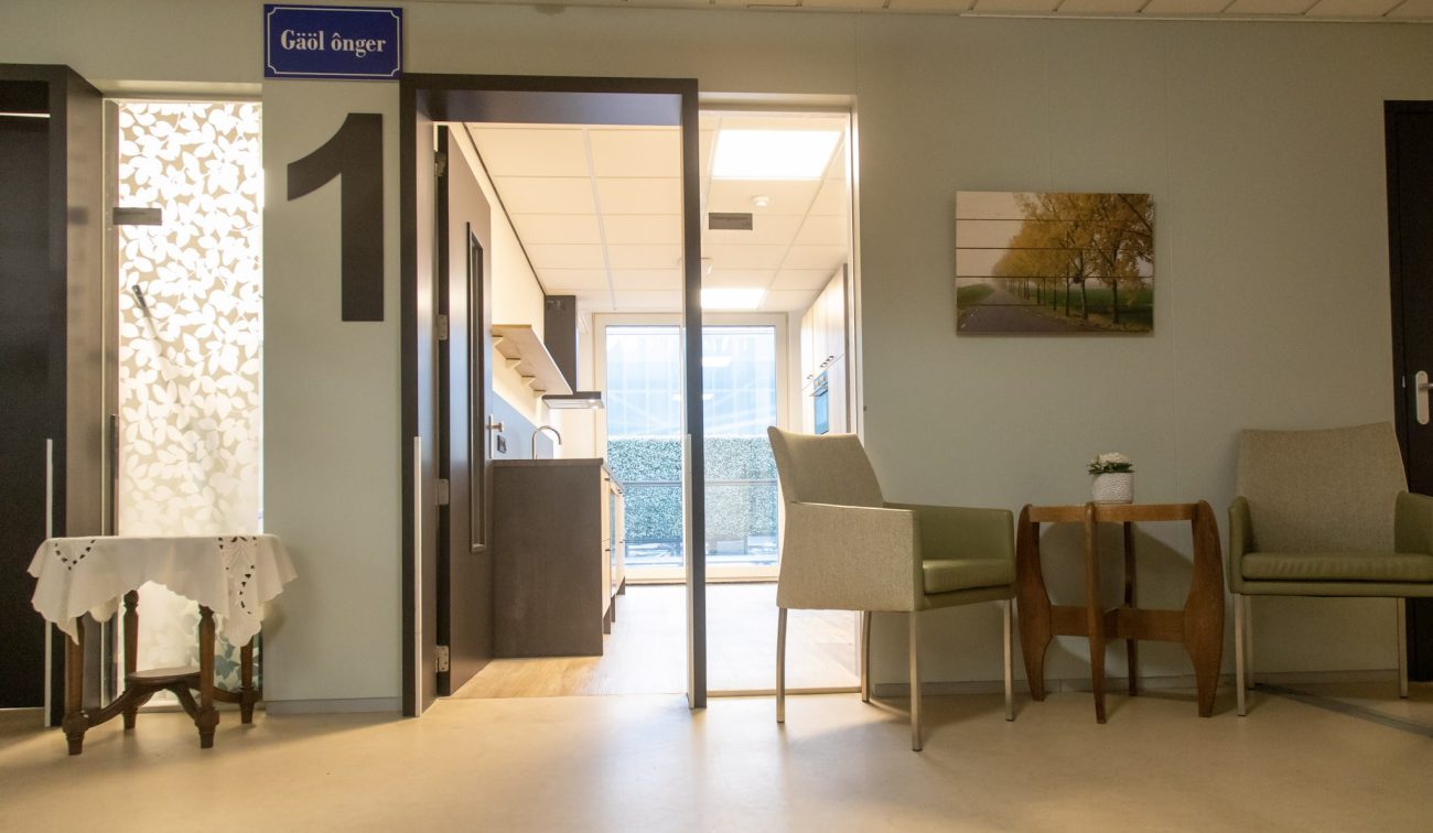 Zithoek in zorgcentrum met twee fauteuils en houten bijzettafel; links deur met groot cijfer 1 naar een lichte kitchenette, aan de muur een foto van een laan.