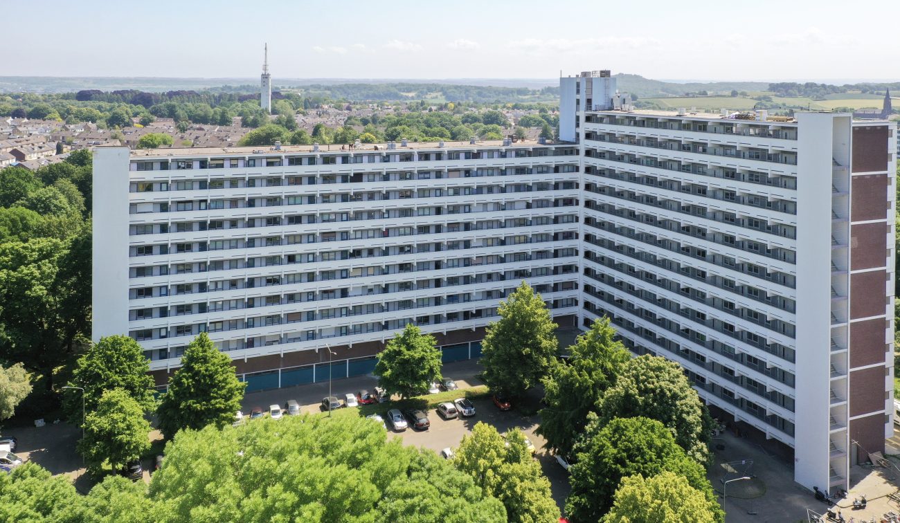 Luchtfoto van L-vormige galerijflat Veliahof in Maastricht, met witte gerenoveerde gevels en lange balkonstroken, omgeven door bomen en parkeerplaatsen.