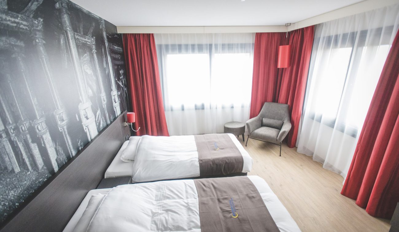 Interieur hotelkamer Geleen – twee bedden, rode overgordijnen, sheer vitrage en grijze stoel met bijzettafel