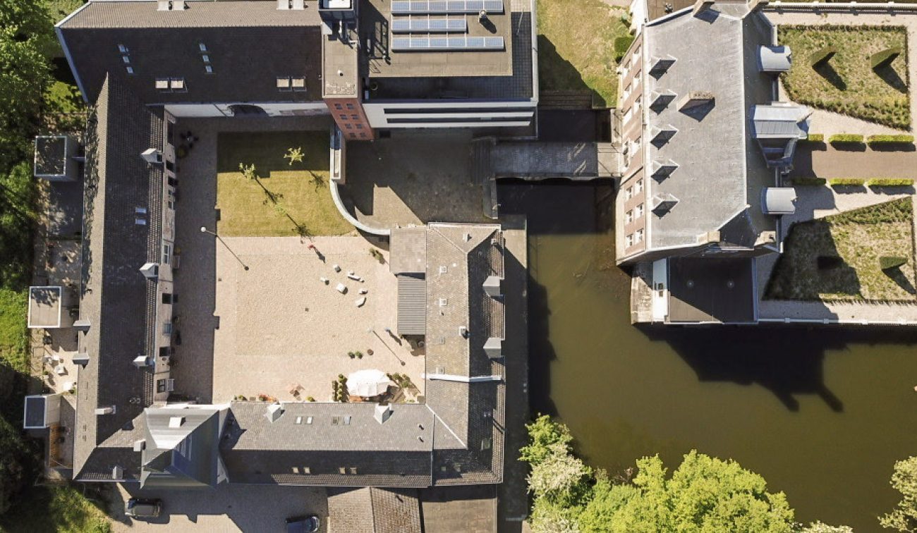 Verticale luchtfoto van carré-hoeve met geplaveide binnenplaats naast een waterloop en brug; combinatie van monument en modern woonblok