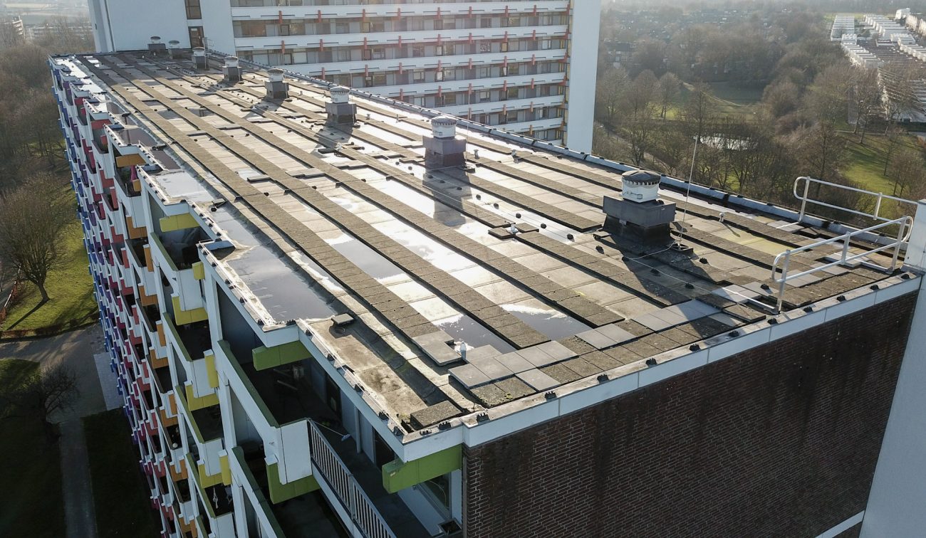 Luchtfoto van het platte dak van een galerijflat met bitumenbanen, ventilatie-units en valbeveiligingsrailing; gekleurde balkonpanelen aan de gevel.
