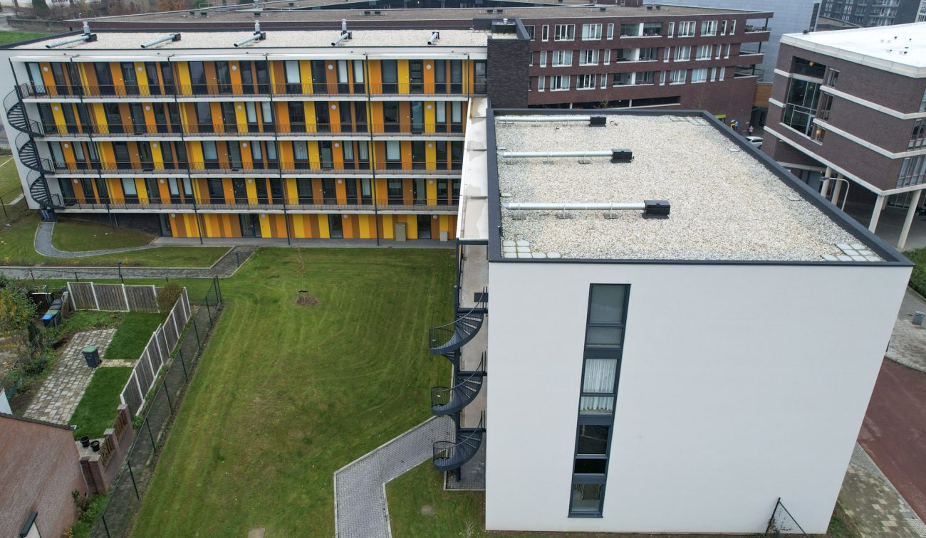 Bovenaanzicht van een binnenhof: rechts een wit appartementengebouw met plat grinddak en spiraaltrappen, links een L-vormig galerijblok met verticale geel-oranje gevelpanelen; daartussen een groot grasveld met paden en hekken.