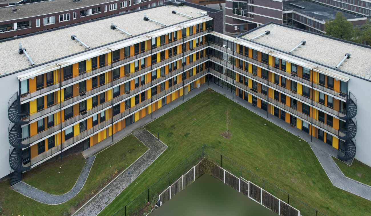 Nieuwbouw met twee vleugels in een L-vorm; galerijen met gele en oranje panelen, metalen balustrades en buitentrappen rond een grasrijke binnentuin.