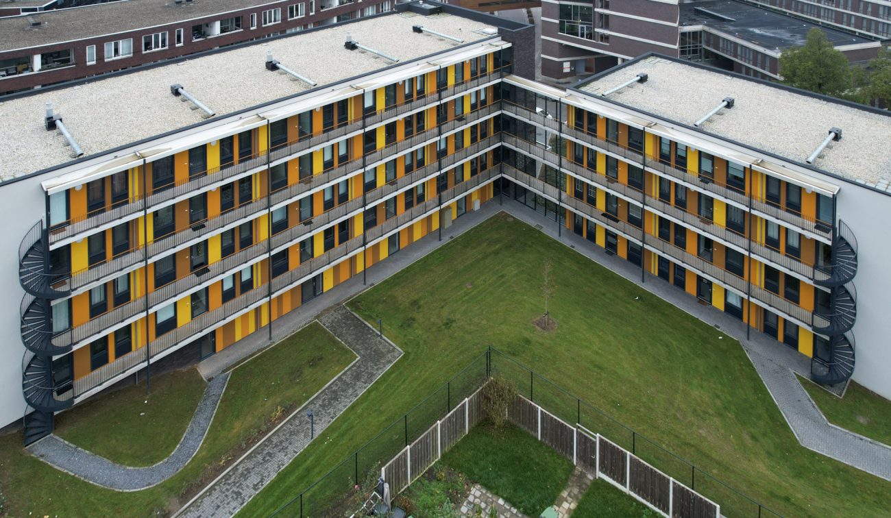 Luchtfoto van nieuwbouw Magistraat Heerlen: U-vormige blokken met doorlopende balkons, gele en oranje panelen en een centrale groene binnentuin.”