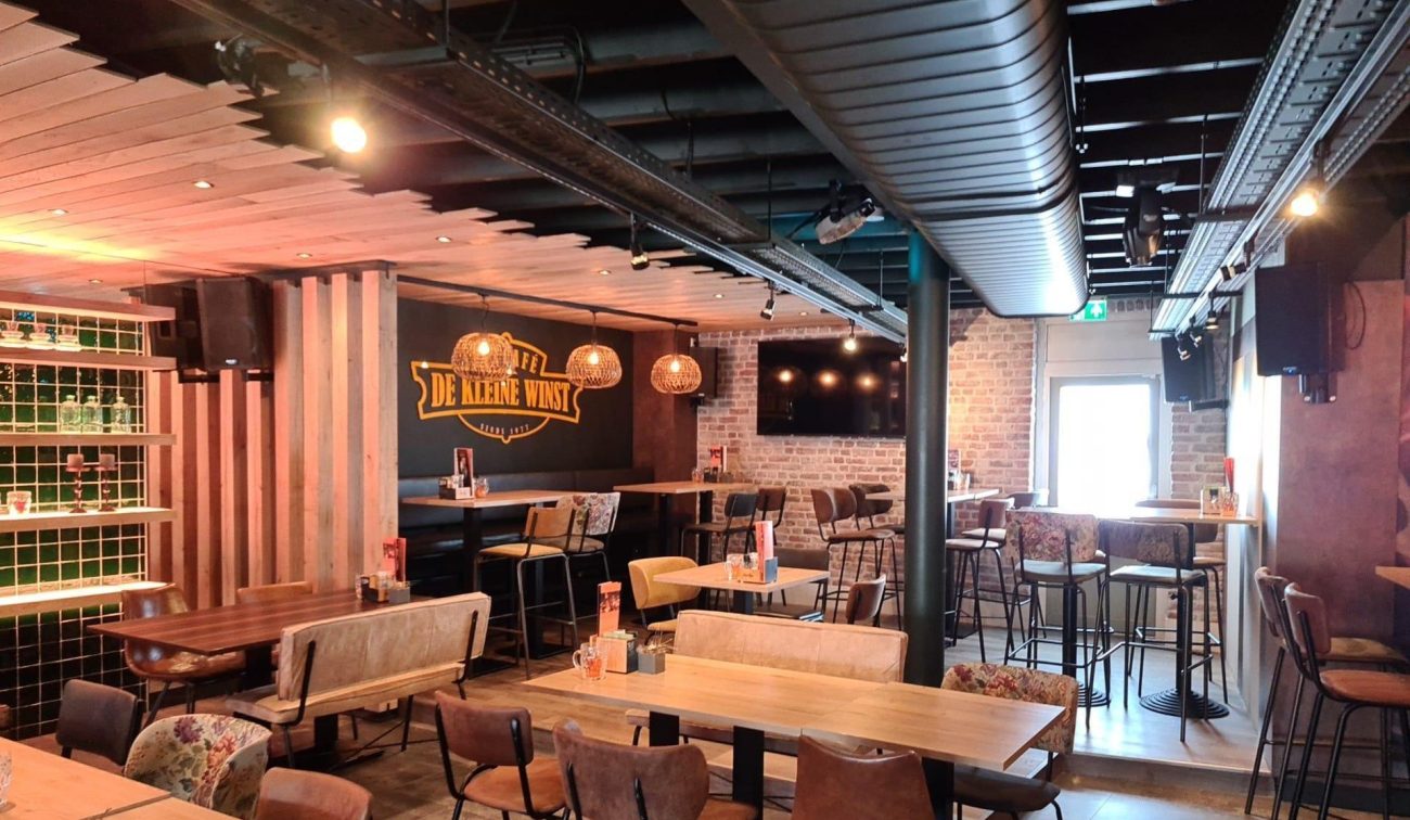 Interieur eetcafé De Aanwinst met industriële look: houten tafels en banken, hoge krukken, bakstenen accentmuur met logo, warme hanglampen en zichtbare luchtkanalen aan het plafond.