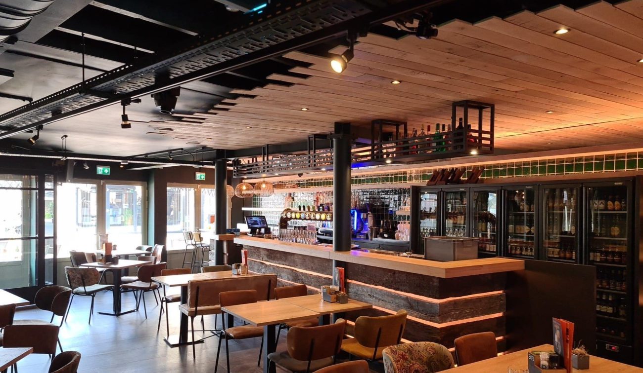 Warm eetcafé-interieur met houten bar, tapkranen en tafels met leren stoelen onder een houten plafond.