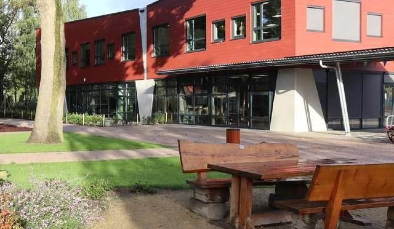 Nieuwbouw van basisschool Den Doelhof in Meijel met rood beklede gevel en grote ramen; voor het gebouw liggen houten picknickbanken op het groene schoolplein.
