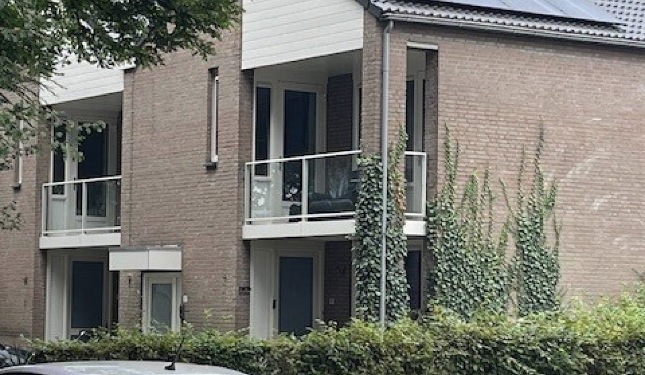 Appartementen/maisonnettes in Stramproy (groot onderhoud) met bakstenen gevel, wit topgeveldeel en balkons met glazen balustrades; op het schuine dak liggen zonnepanelen. Voor het gebouw staan een haag en een geparkeerde bedrijfsauto.